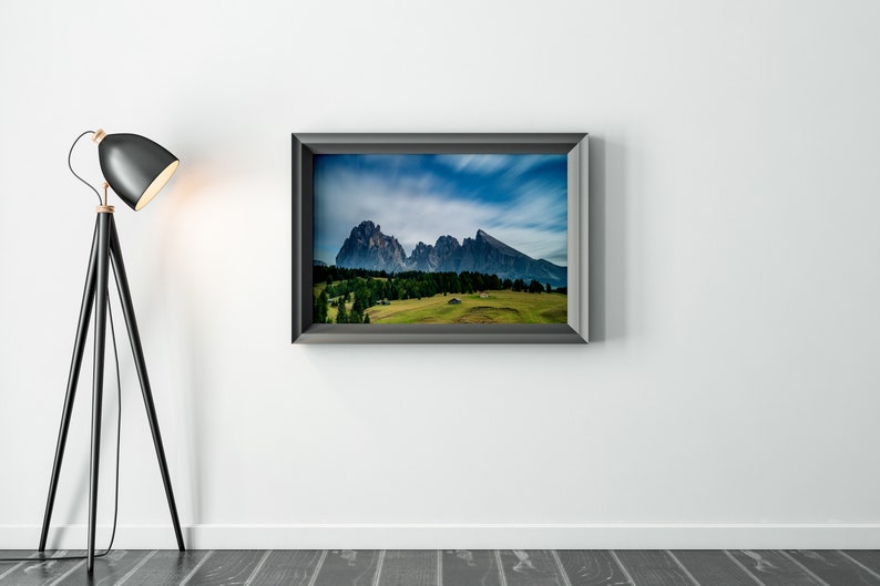 May include: A framed landscape photograph of a mountain range with a green valley and a blue sky. A black floor lamp with a black lampshade is to the left of the framed photograph.