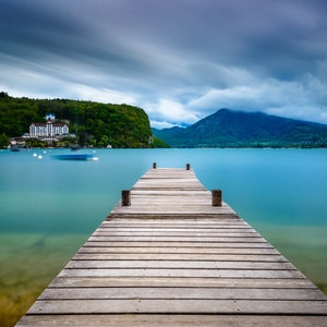 May include: A wooden pier stretches into a tranquil turquoise lake under a cloudy sky. A mountain range and a building on a green hillside are visible in the background. The image has a long exposure effect.