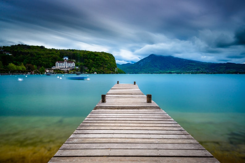 May include: A wooden pier stretches into a tranquil turquoise lake under a cloudy sky. A mountain range and a building on a green hillside are visible in the background. The image has a long exposure effect.