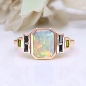 May include: A rose gold ring with a large, rectangular, white opal gemstone. The ring has two black and two green gemstones on either side of the opal.