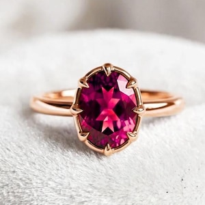 May include: A rose gold ring featuring a central oval-cut deep pink gemstone. The stone is held in place by a prong setting with multiple metal claws that secure the gem along its perimeter, set on a smooth, polished rose gold band.