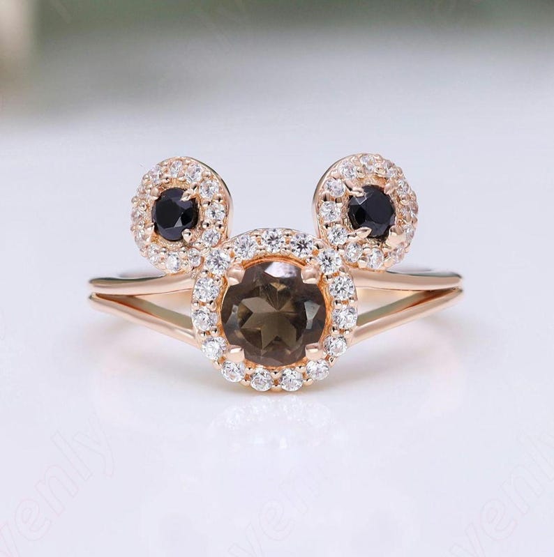 May include: A rose gold ring with a brown gemstone surrounded by smaller white gemstones. The ring has two smaller circles of white gemstones with black gemstones in the center, forming a Mickey Mouse head shape.