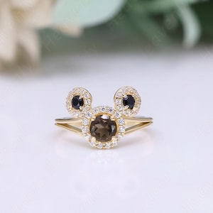 May include: A gold ring with a brown gemstone surrounded by smaller white gemstones. The ring has two black gemstones on either side of the brown gemstone, forming a Mickey Mouse shape.