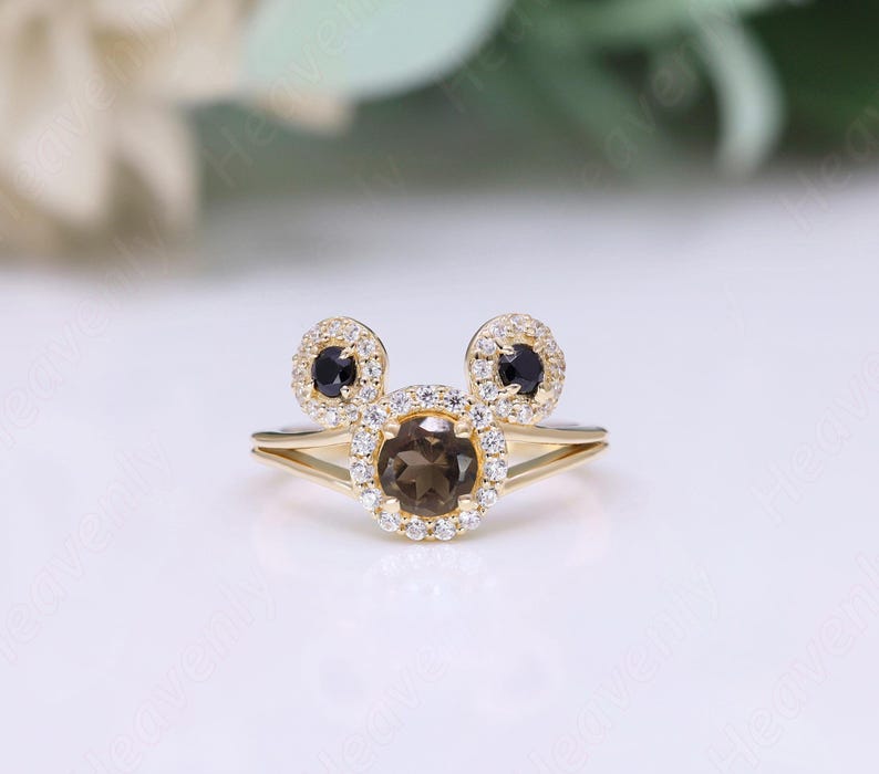 May include: A gold ring with a brown gemstone surrounded by smaller white gemstones. The ring has two black gemstones on either side of the brown gemstone, forming a Mickey Mouse shape.