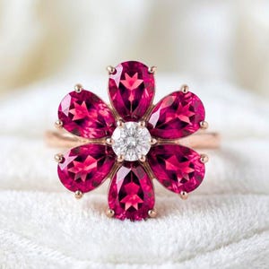 May include: A rose gold ring featuring a floral design with five pear-cut deep pink gemstones arranged as petals around a single round brilliant-cut clear white gemstone center. The band is thin and polished, showcasing a vibrant, symmetrical arrangement.