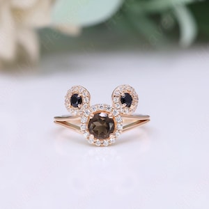 May include: A rose gold ring with a brown gemstone surrounded by smaller clear gemstones. The ring has two smaller black gemstones on either side of the larger gemstone, forming a Mickey Mouse shape.