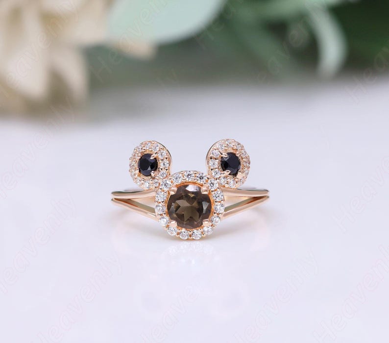 May include: A rose gold ring with a brown gemstone surrounded by smaller clear gemstones. The ring has two smaller black gemstones on either side of the larger gemstone, forming a Mickey Mouse shape.