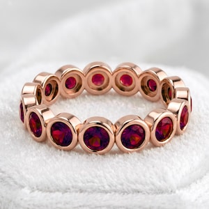 May include: A rose gold eternity band featuring a continuous row of round, faceted, deep red gemstones set in individual circular bezels. The ring displays a uniform design with polished metal edges surrounding each vibrant red stone.