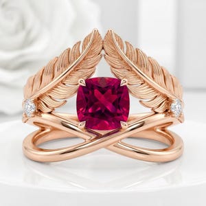 May include: A rose gold ring featuring a central cushion-cut deep pink gemstone. The band is designed with two symmetrical feather motifs extending upwards, each accented with a small round white diamond at the base of the feather structure.