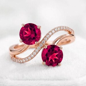 May include: A rose gold bypass ring featuring two round-cut vibrant red gemstones held in four-prong settings. The band is split into two flowing, intertwined curves, with one side encrusted with small, clear pavé-set white stones.