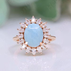 May include: A rose gold ring with a large oval-shaped blue gemstone surrounded by a halo of smaller clear gemstones.