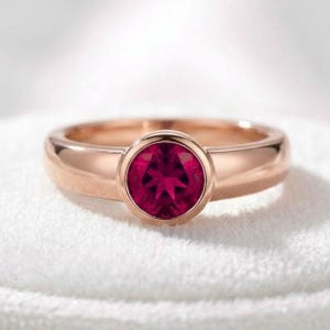 Pode incluir: Um anel de ouro rosa com uma pedra preciosa redonda, facetada e de cor rosa profundo, fixada numa cravação lisa. A banda é polida e sólida, com um perfil arredondado que se estreita ligeiramente em direção à pedra central, criando um visual.
