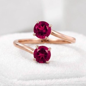 May include: A bypass ring crafted from rose gold, featuring two round-cut, deep red rubies set in four-prong mountings. The open-ended band design allows the two vibrant gemstones to sit prominently at the front, creating a balanced, symmetrical look.