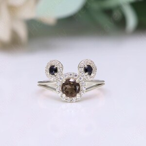 May include: A silver ring with a brown gemstone surrounded by smaller white gemstones. The ring is shaped like Mickey Mouse's head with two black gemstones for the ears.