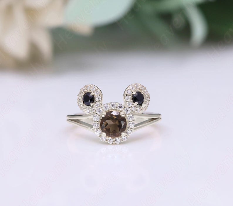 May include: A silver ring with a brown gemstone surrounded by smaller white gemstones. The ring is shaped like Mickey Mouse's head with two black gemstones for the ears.