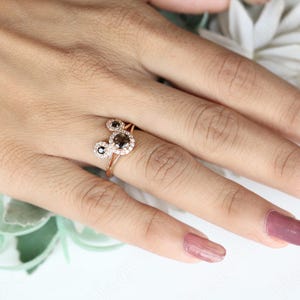 May include: A rose gold ring with two round black diamonds set in a halo of smaller white diamonds. The ring is on a finger with pink nail polish.