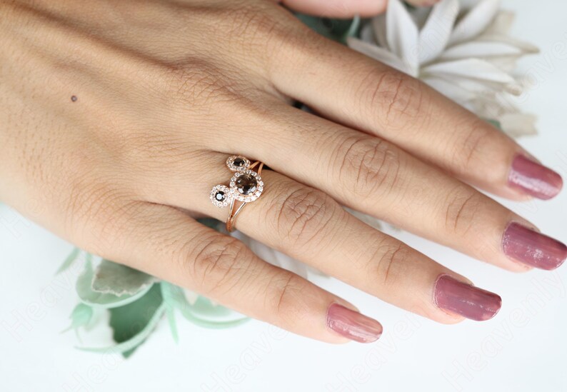May include: A rose gold ring with two round black diamonds set in a halo of smaller white diamonds. The ring is on a finger with pink nail polish.