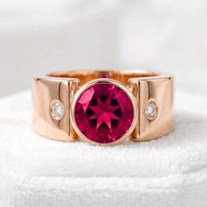 Pode incluir: Um anel de banda larga em ouro rosa com uma grande pedra preciosa redonda e facetada de cor rosa profundo no centro. A banda é acentuada com duas pedras brancas redondas mais pequenas, uma de cada lado da pedra central rosa.
