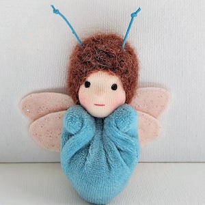 May include: A handcrafted fairy doll with a blue body and brown hair. The doll features pink, glitter-covered wings and a fabric face with black eyes and a red mouth. Blue antennae are attached to the head, giving it a whimsical look.
