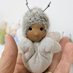 May include: A handmade doll with a grey fuzzy hat and matching wings. The doll has a brown fabric face with black eyes and a small pink mouth. The doll is held in a hand.