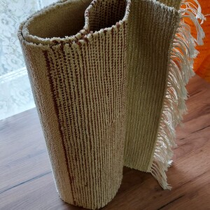 Peut inclure: Un tapis beige et brun &agrave; rayures roul&eacute; avec des franges sur les bords. Le tapis est roul&eacute; et pos&eacute; sur une surface en bois.