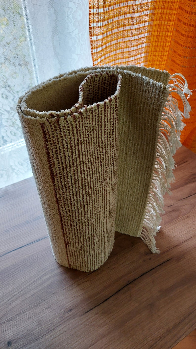 Peut inclure: Un tapis beige et brun &agrave; rayures roul&eacute; avec des franges sur les bords. Le tapis est roul&eacute; et pos&eacute; sur une surface en bois.