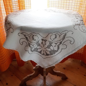 May include: White tablecloth with a delicate black embroidered floral design. The tablecloth is draped over a wooden table with four legs.