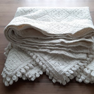 May include: A white crocheted throw blanket with a delicate lace pattern and fringe trim. The blanket is folded and displayed on a wooden surface.