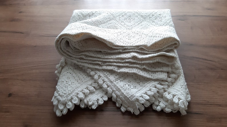 May include: A white crocheted throw blanket with a delicate lace pattern and fringe trim. The blanket is folded and displayed on a wooden surface.