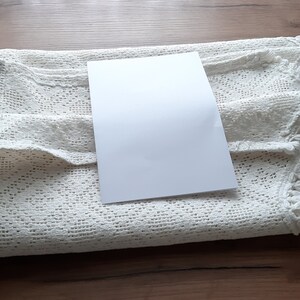 May include: A white crocheted tablecloth with a white card on top. The tablecloth has a delicate lace pattern and a scalloped edge.
