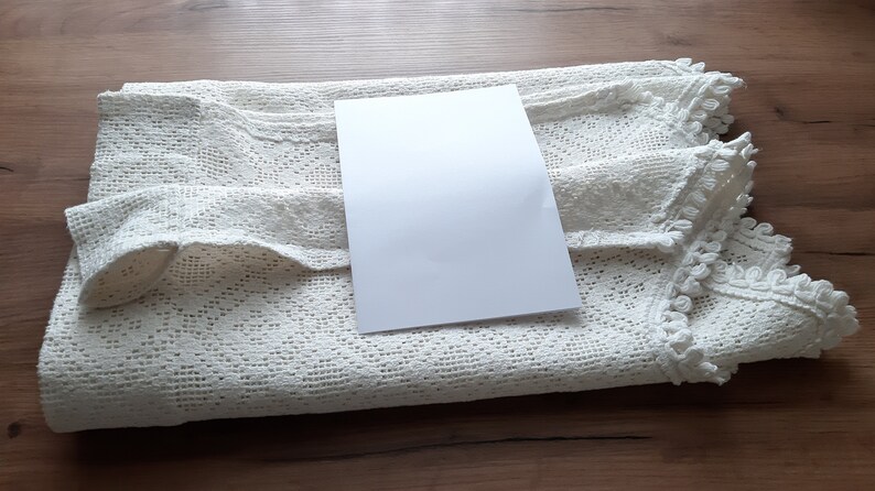 May include: A white crocheted tablecloth with a white card on top. The tablecloth has a delicate lace pattern and a scalloped edge.