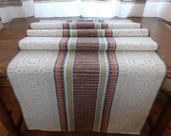 May include: A white table runner with a brown, green, blue, and orange striped design. The runner is folded in thirds and is lying on a wooden table.