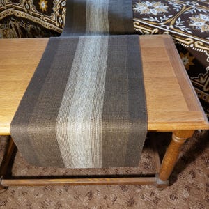 May include: A brown and gray striped table runner is draped over a wooden table. The runner features vertical stripes in varying shades of brown and gray. The table has a light wood finish and is supported by turned legs.