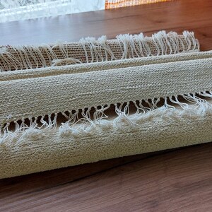 Peut inclure: Un tapis beige roul&eacute; avec des franges blanches sur les bords. Le tapis est fait d'un mat&eacute;riau tiss&eacute; et a un aspect naturel et rustique.