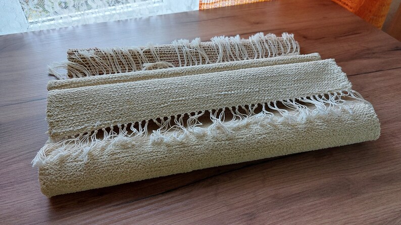 Peut inclure: Un tapis beige roul&eacute; avec des franges blanches sur les bords. Le tapis est fait d'un mat&eacute;riau tiss&eacute; et a un aspect naturel et rustique.