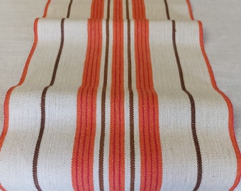 May include: A white table runner with orange and brown stripes. The runner is made of a woven fabric and has a slightly textured surface.