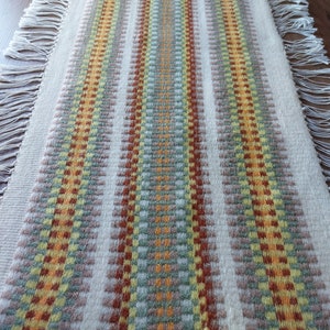 May include: A white table runner with a woven pattern of orange, yellow, and green stripes. The runner has fringe on both ends.