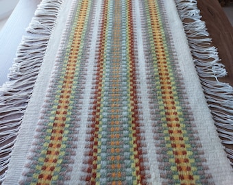 May include: A white table runner with a woven pattern of orange, yellow, and green stripes. The runner has fringe on both ends.