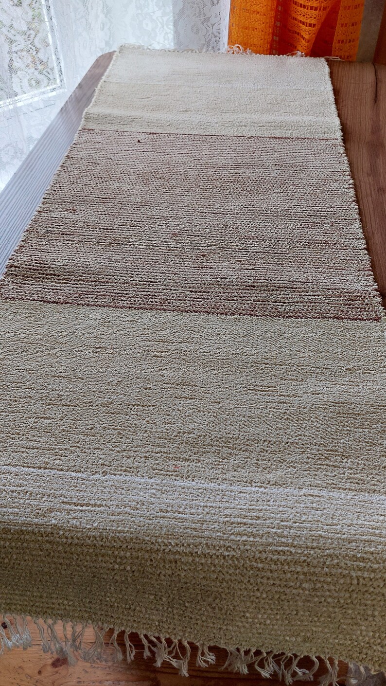 Peut inclure: Un tapis tiss&eacute; de couleur beige naturel avec une bande brune au milieu. Le tapis a une frange sur le bord inf&eacute;rieur.