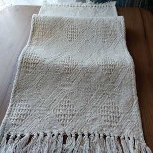 Op de afbeelding: Een witte gehaakte tafelloper met een ruitpatroon en franjes.