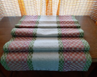 May include: A white, green, and pink checkered table runner with a woven design. The runner is folded in thirds and is lying on a wooden table.