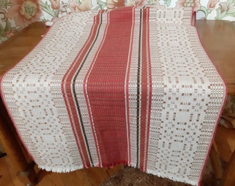 May include: A white table runner with red and black stripes and a woven pattern. The runner has a fringed edge.