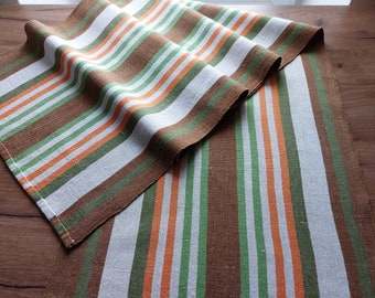 May include: A brown, white, green, and orange striped table runner. The runner is made of a woven fabric and has a slightly textured surface.