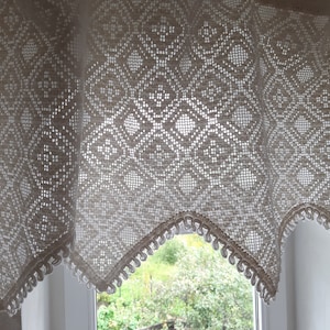 May include: A white crocheted valance with a diamond pattern and scalloped edges hangs over a window with a view of green trees.