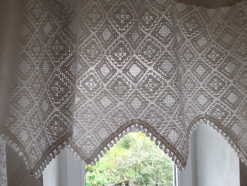 May include: A white crocheted valance with a diamond pattern and scalloped edges hangs over a window with a view of green trees.
