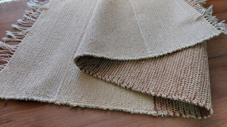 Peut inclure: Un tapis tiss&eacute; beige et brun avec des franges sur les bords. Le tapis est pli&eacute; en deux, r&eacute;v&eacute;lant le motif tiss&eacute; brun en dessous.
