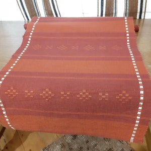 May include: A red and white striped table runner with a woven pattern. The runner has a white border with a small diamond pattern.