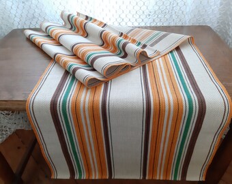 May include: A striped table runner with orange, brown and green stripes on a white background. The runner is folded in half and laid on a wooden table.