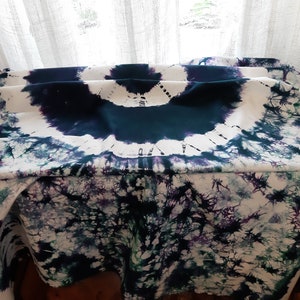 May include: A white fabric tablecloth with a tie-dye pattern in shades of blue, green, and purple. The fabric is folded in the center, creating a circular design.