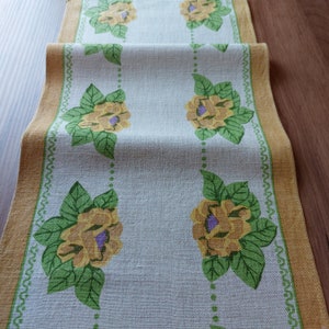 May include: A white table runner with a yellow and green floral pattern. The runner has a gold border with a decorative pattern.